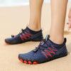 Person trägt Navy/blaue Barfußschuhe mit roten Akzenten am Sandstrand. Die Schuhe haben eine flexible, zehenförmige Sohle und elastische Schnürung.