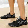Person trägt schwarze Barfußschuhe mit grauen Akzenten am Sandstrand. Die Schuhe haben eine flexible, zehenförmige Sohle und elastische Schnürung.