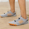 Person trägt graue Barfußschuhe mit orangenen Akzenten am Sandstrand. Die Schuhe haben eine flexible, zehenförmige Sohle und elastische Schnürung.